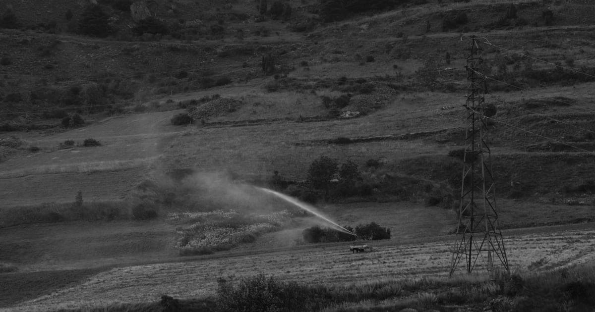 Riego por aspersión lanzando un chorro de agua sobre un campo en un paisaje árido y montañoso, en blanco y negro.